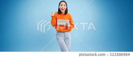 Amazed korean woman with digital tablet, looks surprised and excited, standing over blue background 122886984