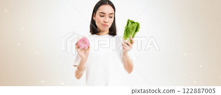 Healthy lifestyle concept. Young woman deciding between healthy vegetables, cabbage and delicious glazed doughnuts, white background 122887005
