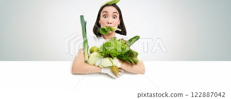 Close up portrait of asian girl munching lettuce leaf, holding green oranic vegetables, leads healthy lifestyle with nutritious food, white background Close up portrait of asian girl munching lettuce leaf, holding green oranic vegetables, leads healthy lifestyle with nutritious food, white background 122887042