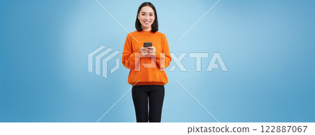 Girl with smartphone in hands smiling, using mobile application on her phone, sending message or order something online, blue background Girl with smartphone in hands smiling, using mobile application on her phone, sending message or order something online, blue background 122887067
