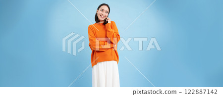 Korean girl in orange sweater talks on mobile phone, speaks on cellphone with happy smiling face, orders delivery, consults with someone on telephone 122887142