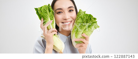 Healthy food and vegan lifestyle. Close up portrait of smiling asian woman, looks happy, shows her face with lettuce, eats cabbage, white background 122887143