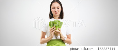 Healthy diet and raw food. Sad and unhappy asian woman, holding fresh lettuce leaves, cabbage in hands, eating vegetables, white background 122887144