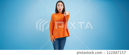 Amazed korean girl, points finger at camera, drops jaw from surprise, looks impressed at smth, stands over blue background 122887157