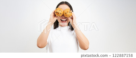 Close up portrait of funny asian girl, holds cupcakes near face, makes happy faces, white studio background Close up portrait of funny asian girl, holds cupcakes near face, makes happy faces, white studio background 122887158