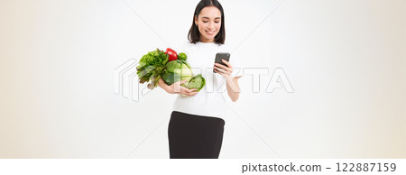 Portrait of korean woman holds vegetables, orders green organic food on smartphone app, white background Portrait of korean woman holds vegetables, orders green organic food on smartphone app, white background 122887159
