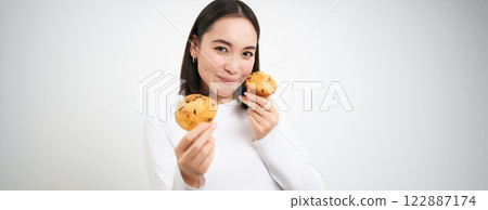 Image of korean woman enjoys eating baked pastry, showing two tasty cupcakes near face and smiles, white background 122887174