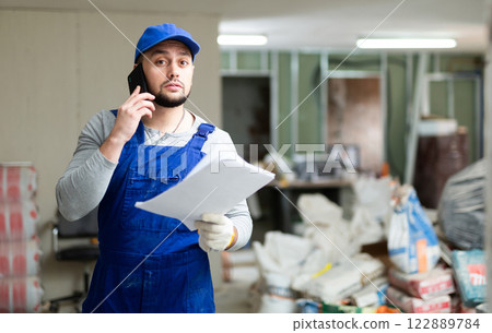 Focused bearded contractor in blue overall standing at construction site indoors, discussing work process on phone 122889784