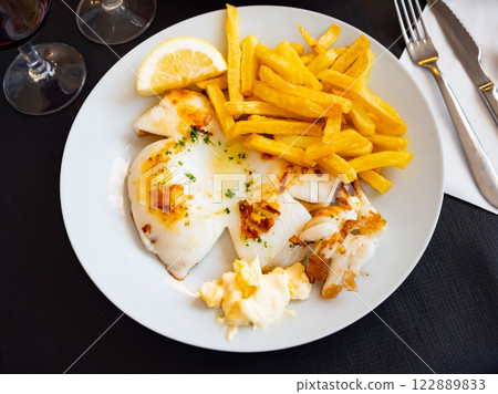 fried cuttlefish drizzled with white garlic and parsley sauce,lemon wedge and french fries on plate 122889833