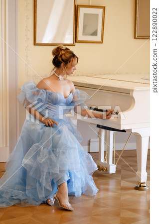 Elegant woman in a light blue ball gown in a luxurious classic interior plays a white piano 122890022