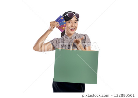 Portrait of Asian Indonesian woman with sunglasses holding credit cards while carrying shopping bag isolated over white background. Copy space or mockup. Concept of shopping, lifestyle, or consumerism 122890501