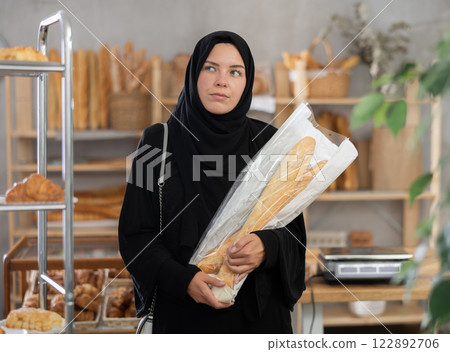 Muslim woman buyer with baguettes in plastic package stands in trading floor of bakery 122892706