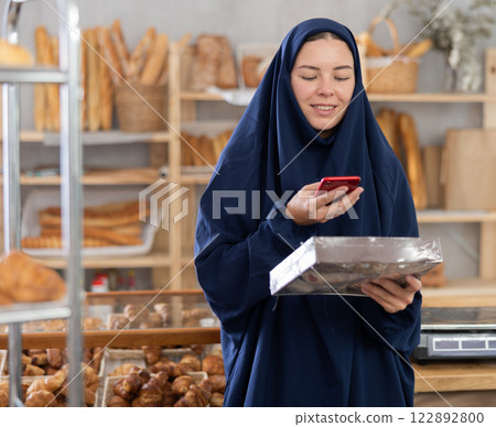 European Muslim woman in hijab customer at bakery scans QR code on cookie package using phone camera European Muslim woman in hijab customer at bakery scans QR code on cookie package using phone camera 122892800