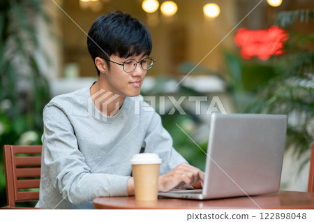 A young Asian man works remotely on his laptop from a coffee shop, sitting at an outdoor table. 122898048