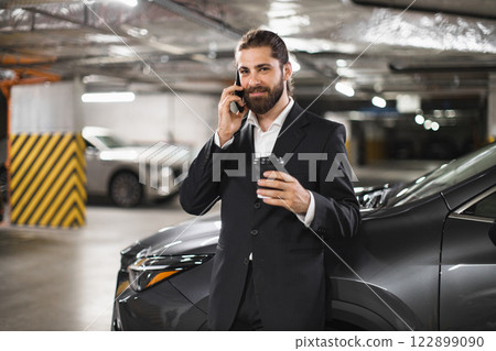 Confident Caucasian male businessman in suit talks on phone with coffee cup in hand, standing in car park. Professional and focused demeanor suggests important business conversation. 122899090