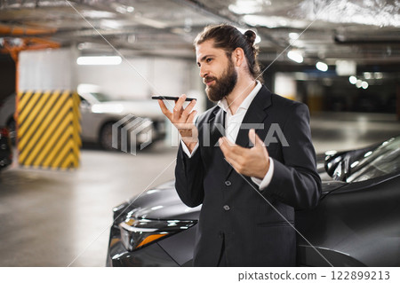 Confident Caucasian male in suit recording voice message on smartphone in parking garage. Business communication and technology concept. Middle-aged professional multitasking in urban setting. Confident Caucasian male in suit recording voice message on smartphone in parking garage. Business communication and technology concept. Middle-aged professional multitasking in urban setting. 122899213