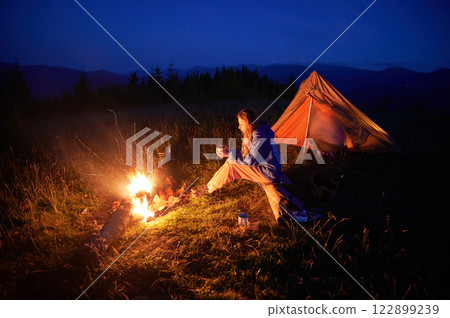 Woman hiker sits by campfire near her illuminated tent in soft glow of twilight, surrounded by serene mountain scenery. Clear night sky above adds peaceful ambiance to quiet wilderness setting. 122899239
