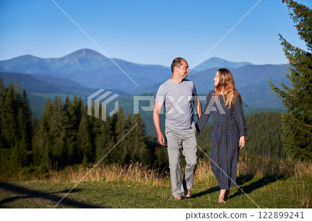 Couple holds hands, walk barefoot through meadow, enjoying stunning mountain view. Lush green forest and distant mountains under clear blue sky create serene and romantic atmosphere. 122899241