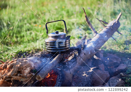 Close-up of metal kettle heating over campfire. Flames and smoke curl around well-used kettle, which rests on logs. Blur of green grass, emphasizing rustic outdoor setting on background. 122899242