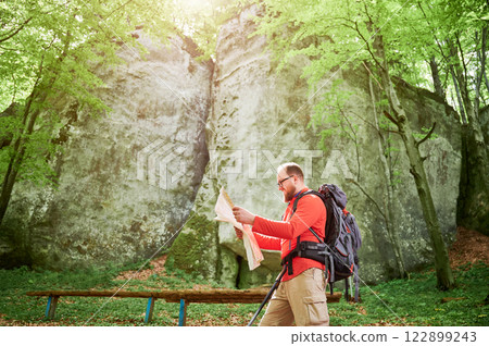 Happy bearded tourist man studies map while stands in front of large rock in dense forest. Smiling traveler with grey backpack and glasses, planning outdoor adventure, hiking or climbing route. Happy bearded tourist man studies map while stands in front of large rock in dense forest. Smiling traveler with grey backpack and glasses, planning outdoor adventure, hiking or climbing route. 122899243