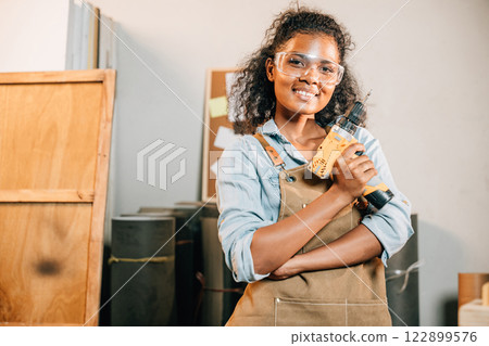 Smiling carpenter woman holding a power drill in a workshop, with a wooden stool and small house models nearby. Surrounded by tools, showcasing her craft and enthusiasm for woodworking projects 122899576