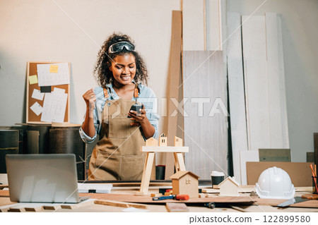 Carpenter woman in a workshop uses her smartphone. Business owner and product designer managing projects online, reviewing designs, and gathering feedback for evaluation and improvement Carpenter woman in a workshop uses her smartphone. Business owner and product designer managing projects online, reviewing designs, and gathering feedback for evaluation and improvement 122899580
