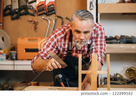 Senior carpenter uses a ruler and set square to measure and assemble a wooden structure with precise alignment, focusing on craftsmanship. Workshop is organized with tools and materials Senior carpenter uses a ruler and set square to measure and assemble a wooden structure with precise alignment, focusing on craftsmanship. Workshop is organized with tools and materials 122899582