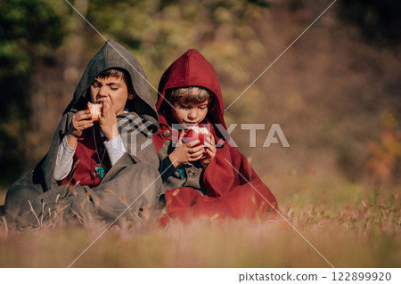 Hobbits boys in costumes sitting on autumn ground to eat apples. Fantasy journey, halloween 122899920