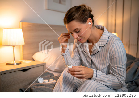 Woman inserts earplugs before going to sleep by nightlight while sitting in her pajamas on her bed. Sleep hygiene. Remedies for insomnia Woman inserts earplugs before going to sleep by nightlight while sitting in her pajamas on her bed. Sleep hygiene. Remedies for insomnia 122902289