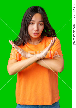 Young woman in orange shirt making a cross gesture against a bright green background 122902332