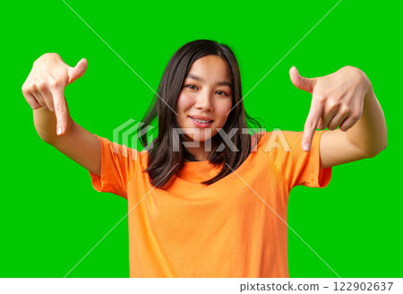 Young woman in orange shirt gestures playfully against bright green backdrop while smiling and having fun Young woman in orange shirt gestures playfully against bright green backdrop while smiling and having fun 122902637