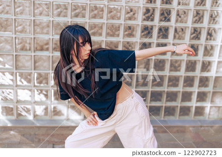 Young woman showcases dance moves outdoors near glass brick wall during sunny day Young woman showcases dance moves outdoors near glass brick wall during sunny day 122902723