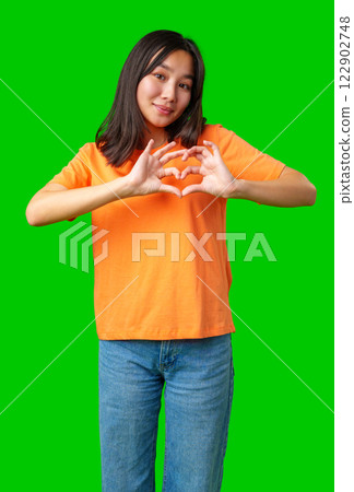 Young person makes heart shape with hands against bright green background during casual indoor setting 122902748