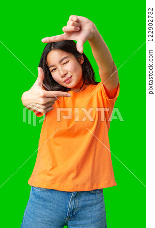 Young woman poses playfully with hand gestures against a bright green background in a casual setting 122902782