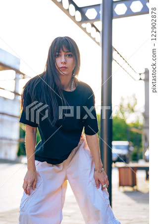 Young woman poses confidently outdoors in urban setting during sunset, showcasing casual fashion style with relaxed demeanor and strong presence 122902878