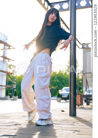 Young woman dancing outdoors in urban setting during sunset wearing casual streetwear 122903029