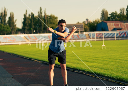 Young man stretches on the athletic track in a sunny stadium during evening practice session Young man stretches on the athletic track in a sunny stadium during evening practice session 122903067