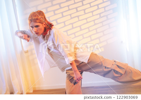 Young model in casual streetwear poses dramatically in a studio with soft lighting and textured wall backdrop Young model in casual streetwear poses dramatically in a studio with soft lighting and textured wall backdrop 122903069