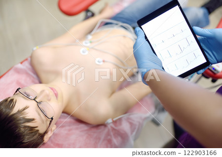 Child under ECG test with doctor. Boy patient lying in medical bed with vacuum sensors. A nurse performs an electrocardiogram of the heart in office of a medical clinic. 122903166
