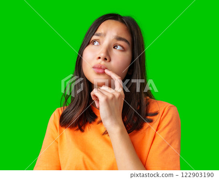 Young woman in bright orange shirt on green background appears thoughtful while pondering a question Young woman in bright orange shirt on green background appears thoughtful while pondering a question 122903190