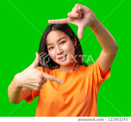 Young woman gestures a frame with her hands against a bright green background while wearing an orange shirt Young woman gestures a frame with her hands against a bright green background while wearing an orange shirt 122903243