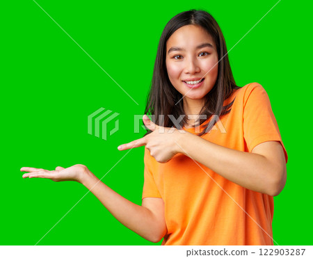 Smiling woman in orange shirt gestures towards an empty space on a bright green background 122903287