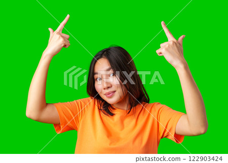 Young woman in orange shirt making playful gestures against a green backdrop in a lively indoor setting 122903424