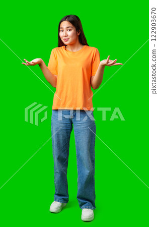 Young woman in an orange shirt gestures with uncertainty against a green background in a playful expression 122903670