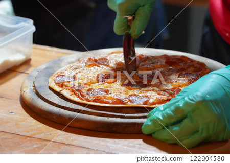 A person wearing green gloves is cutting thin-crust, small-size pizzas on a wooden tray with a pizza cutter. A person wearing green gloves is cutting thin-crust, small-size pizzas on a wooden tray with a pizza cutter. 122904580