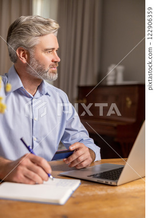 Gray-haired mature man sitting at the laptop and making notes while working Gray-haired mature man sitting at the laptop and making notes while working 122906359