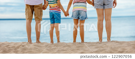 A family stands by holding hands on the seashore at the water's edge looking into the distance. A family stands by holding hands on the seashore at the water's edge looking into the distance. 122907056