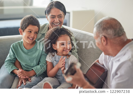 Grandfather playing the guitar for grandmother and children sitting on the sofa in their home. Retired grandparents entertain happy and smiling grandchildren with music on acoustic guitar and bonding 122907238
