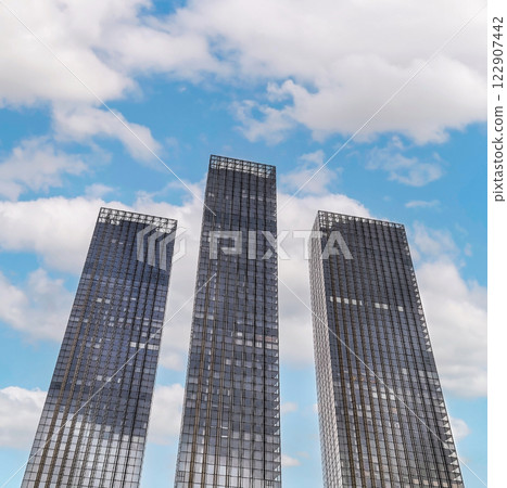 Three modern skyscrapers glisten under a bright blue sky with fluffy clouds during daytime in a bustling urban area Three modern skyscrapers glisten under a bright blue sky with fluffy clouds during daytime in a bustling urban area 122907442