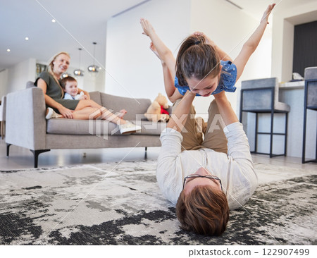 Father with girl on carpet floor and mother relax with baby on sofa in home living room for family love, care and wellness. Happy kid play fun youth game with dad and parents bonding in house lounge 122907499